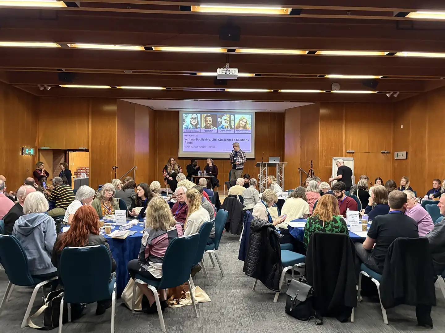 Audience seated at round tables in a wood-panelled conference hall during the Indie Author Lab 'Writing, Publishing, Life: Challenges & Solutions' panel